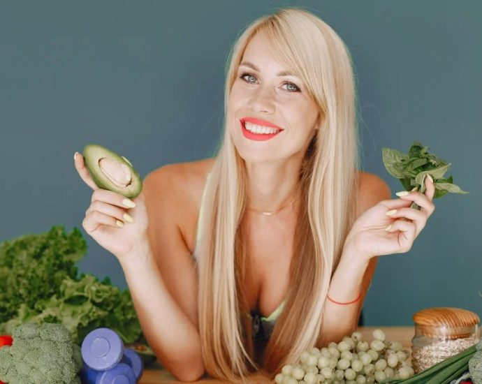 Blonde woman promotes healthy eating with fresh vegetables and fruits on display.